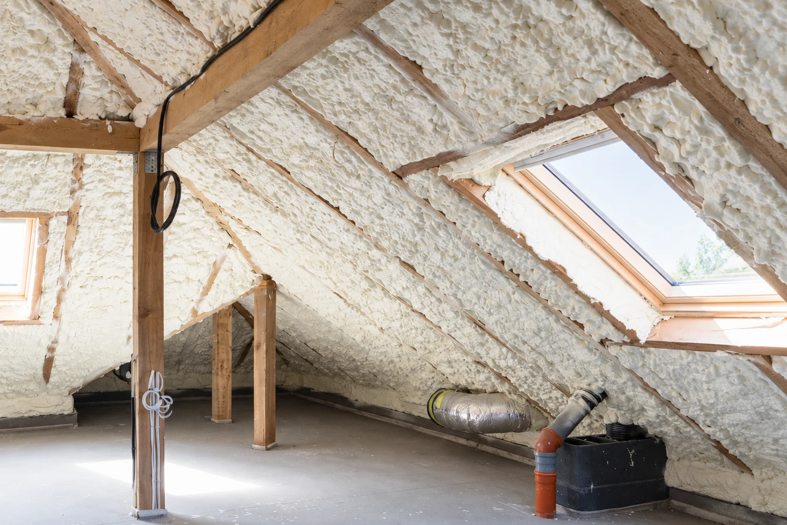 Attic roof and rafters coated with white spray foam insulation, with exposed framing, windows and ductwork.
