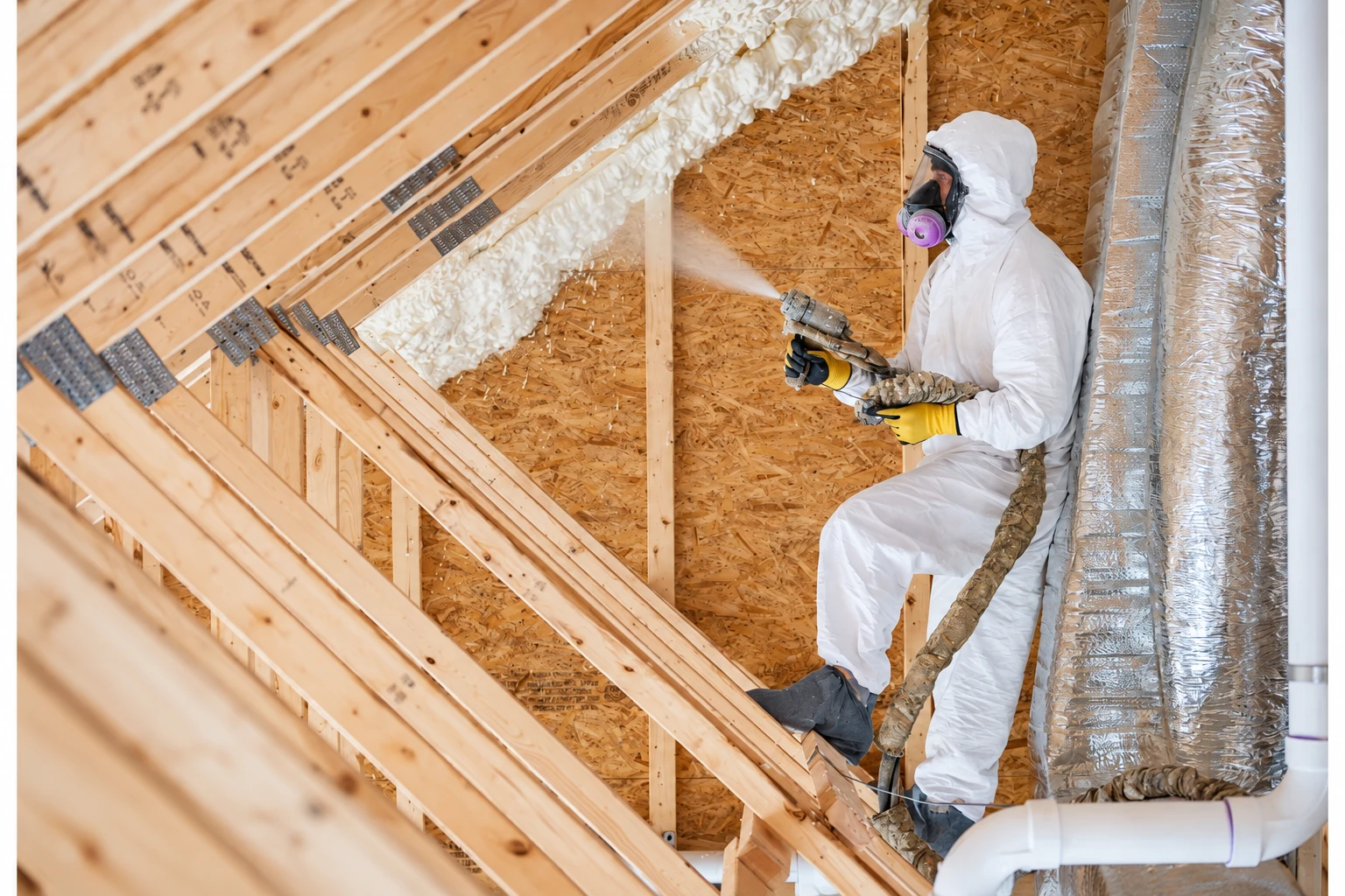 A worker in protective coveralls sprays foam insulation along the rafters of an unfinished attic.