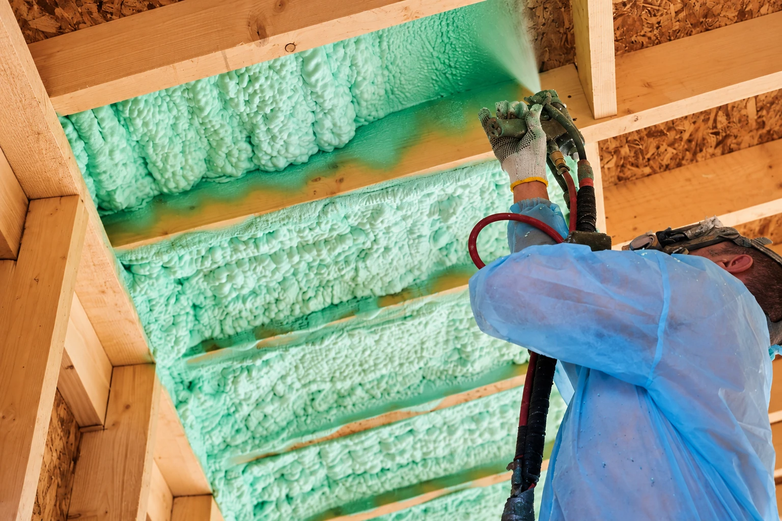 A worker sprays green foam insulation into the spaces between exposed wooden attic rafters.