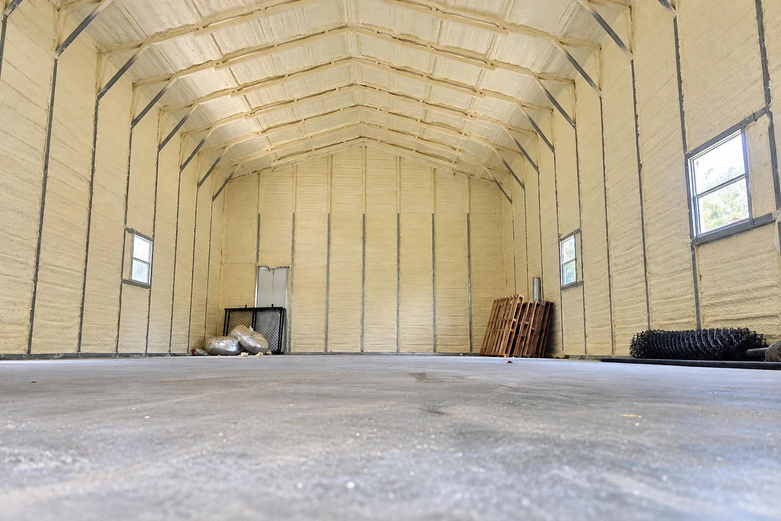 A large arched commercial building has pale spray foam covering its walls and ceiling, with windows and a bare concrete floor.