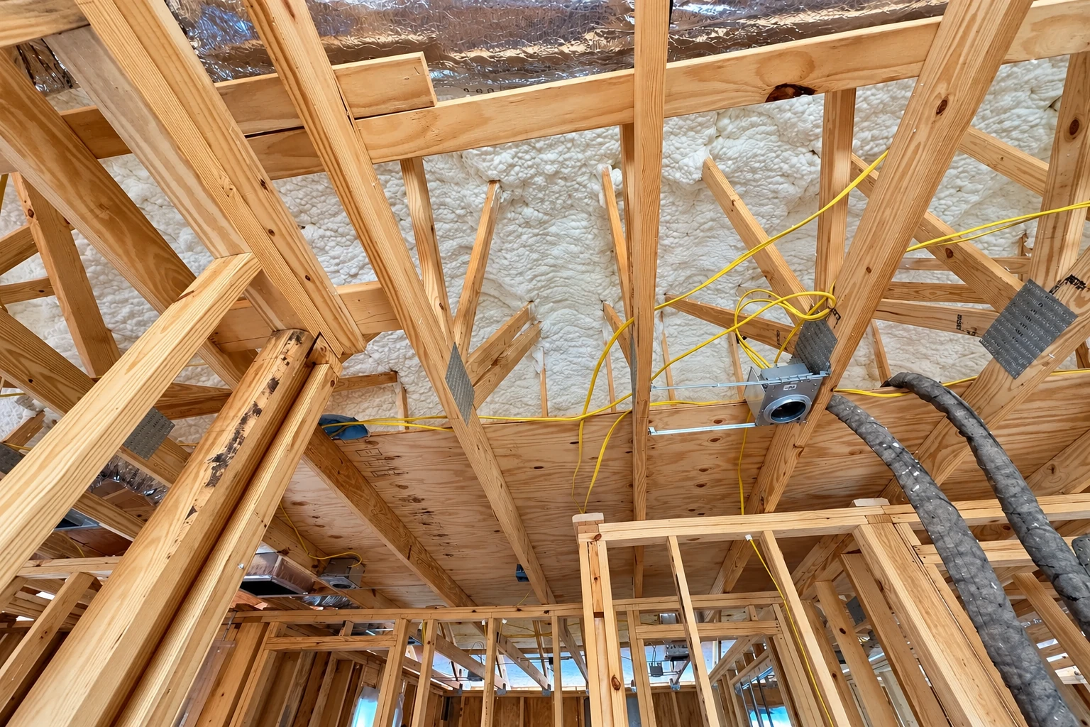 Exposed wooden roof trusses are filled with white spray foam insulation, with cables and ductwork visible.