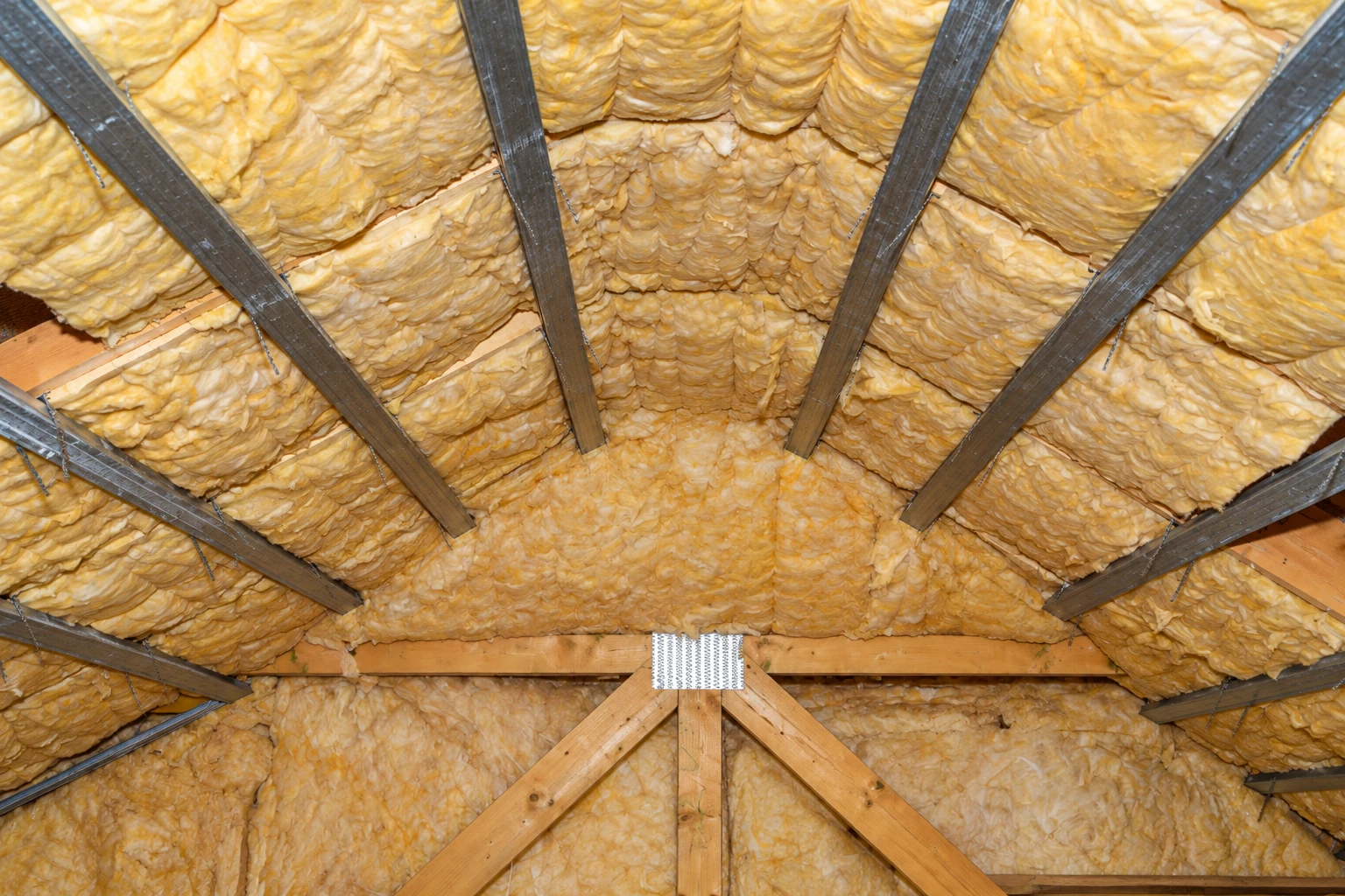 Yellow fiberglass insulation fills the roof rafters and wooden trusses in an attic.